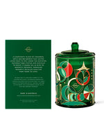 A decorative candle in a cylindrical container with a vibrant green background and an intricate pattern of red, white, and gold geometric shapes. The container has a matching green lid with a knob on top. To the left of the candle is its packaging, featuring the same green color and detailed with text describing the candle’s characteristics.