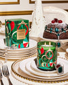 An elegantly set dining table featuring a green and red Glasshouse “Under the Mistletoe” candle and matching packaging box. The table is adorned with white plates, silver cutlery, pearl necklaces as decor, and a chocolate cake decorated with jewels. A white chair with a heart-shaped backrest is partially visible in the background, adding to the festive and luxurious ambiance of the setting.