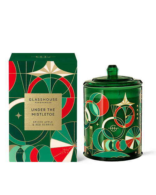 A decorative candle next to its packaging. The candle is housed in a green glass container with a lid, adorned with a pattern of red and gold circles and stars. The packaging box has a matching design and color scheme, featuring the text “Glasshouse Fragrances” at the top, followed by “Under the Mistletoe” in bold letters, and “Spiced Apple & Red Berries” at the bottom indicating the scent. 