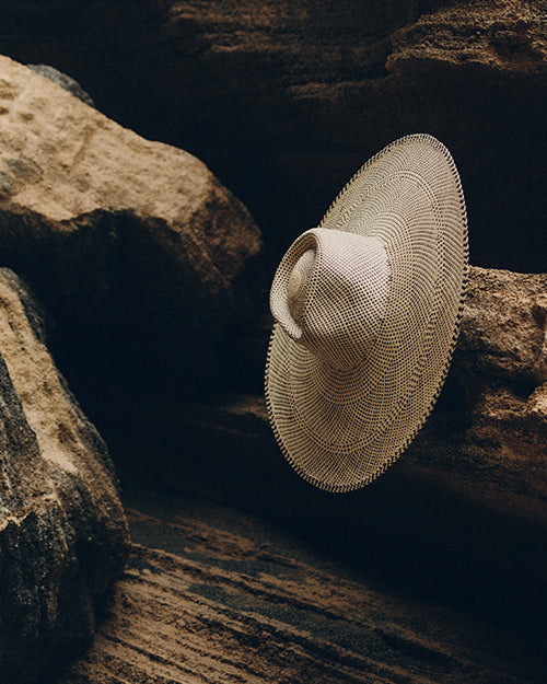 A wide-brimmed hat features a black and white weave pattern that creates a optical illusion effect. The hat has a prominent crown in the center. The hat is pictured elegantly against beach rocks.