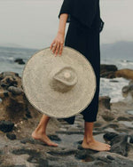 A model holding a wide-brimmed hat features a black and white weave pattern that creates a optical illusion effect. The hat has a prominent crown in the center.
