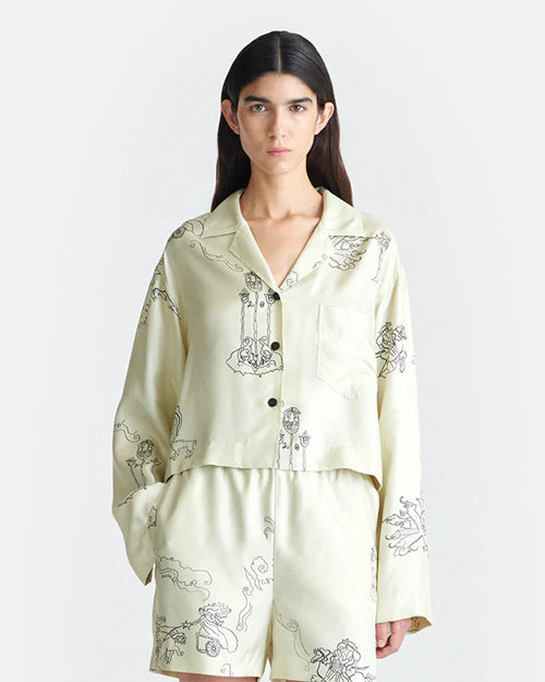 A person is seen wearing a matching pajama set consisting of a long-sleeve button-up shirt and shorts. The pajama set is off-white with black line drawings of various A person is seen wearing a matching pajama set consisting of a long-sleeve button-up shirt and pants. The pajama set is off-white with black line drawings of various whimsical characters and scenes such as warriors on horses and towers. The background is plain white. 