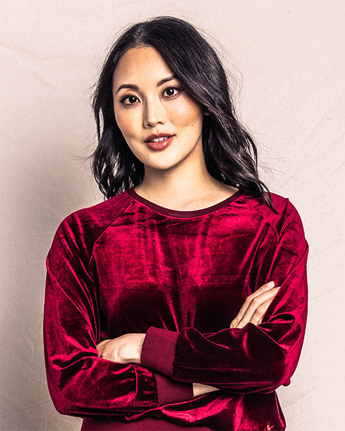 A person wearing a red velvet top with long sleeves . The individual is posed with crossed arms against a neutral-toned backdrop.