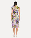 A backside view of a person wearing a sleeveless, knee-length dress with a colorful abstract pattern in shades of purple, green, yellow, and white. The dress has a high neckline with ruffled straps and is cinched at the waist with a matching fabric belt. The person is also wearing white open-toe heels. The background is plain and light-colored.