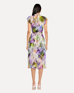 A backside view of a person wearing a sleeveless, knee-length dress with a colorful abstract pattern in shades of purple, green, yellow, and white. The dress has a high neckline with ruffled straps and is cinched at the waist with a matching fabric belt. The person is also wearing white open-toe heels. The background is plain and light-colored.