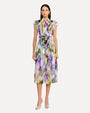 A black-haired model wearing a sleeveless knee-length dress with an colorful abstract pattern, a high neckline with ruffled straps, and is cinched at the waist with a matching fabric belt. The model is also wearing white open-toe heels, standing against a white background.