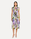 A person wearing a sleeveless, knee-length dress with a colorful abstract pattern in shades of purple, green, yellow, and white. The dress has a high neckline with ruffled straps and is cinched at the waist with a matching fabric belt. The person is also wearing white open-toe heels. The background is plain and light-colored.