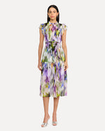 A person wearing a sleeveless, knee-length dress with a colorful abstract pattern in shades of purple, green, yellow, and white. The dress has a high neckline with ruffled straps and is cinched at the waist with a matching fabric belt. The person is also wearing white open-toe heels. The background is plain and light-colored.