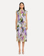 A full-frontal view of a dark-haired model wearing a sleeveless knee-length dress with a colorful abstract pattern, a high neckline with ruffled straps and straight and loosely fitting. The person is also wearing white open-toe heels, standing in front of a plain background.