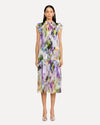 A frontal view of a person wearing a sleeveless, knee-length dress with a colorful abstract pattern in shades of purple, green, yellow, and white. The dress has a high neckline with ruffled straps and straight and loosely fitting. The person is also wearing white open-toe heels. The background is plain and light-colored.