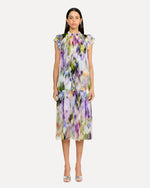 A frontal view of a person wearing a sleeveless, knee-length dress with a colorful abstract pattern in shades of purple, green, yellow, and white. The dress has a high neckline with ruffled straps and straight and loosely fitting. The person is also wearing white open-toe heels. The background is plain and light-colored.