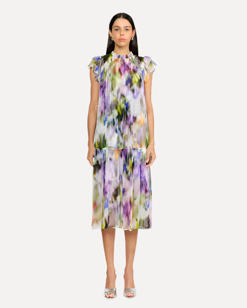 A frontal view of a person wearing a sleeveless, knee-length dress with a colorful abstract pattern in shades of purple, green, yellow, and white. The dress has a high neckline with ruffled straps and straight and loosely fitting. The person is also wearing white open-toe heels. The background is plain and light-colored.