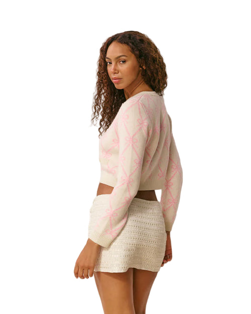 Person standing and looking over one shoulder, wearing a light-colored long-sleeved sweater with a pink pattern and a white textured skirt, against a plain white background.