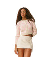 A person wearing a cropped light pink and white cardigan with bow patterns and front buttons, paired with a cream-colored textured mini skirt against a plain white background.