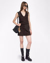A slightly angled full body view of a brunette model wearing a matching set including a brown V-neck tank-top with white rhinestone designs at the shoulders, and a skirt with two pockets in front. She is also wearing black sunglasses, a black purse with multiple charms hanging from it, and black chunky boots. The background is plain white. 