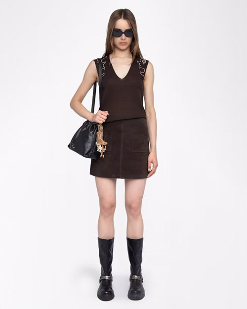 A full body frontal view of a brunette model wearing a matching set including a brown V-neck tank-top with white rhinestone designs shoulders, and a skirt with two pockets in front. She is also wearing black sunglasses, a black purse with multiple charms hanging from it, and black chunky boots. The background is plain white.
