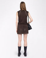 A full body backside view of a brunette model wearing a matching set including a brown tank-top with white rhinestone details at the shoulders, and a short skirt. She is also a black purse with multiple charms hanging from it, and black chunky boots. The background is plain white.