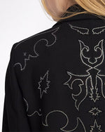 A close-up backside view of a model's shoulder adorned with a black jacket featuring an intricate white embroidery on the back and black pants. 