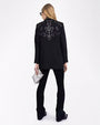 A full body backside view of a blonde model with her hair swept to the side wearing a black jacket with intricate Western style white embroidery at the top of the back and black pants to match. She is also wearing a silver clutch and black boots. The background is plain white.