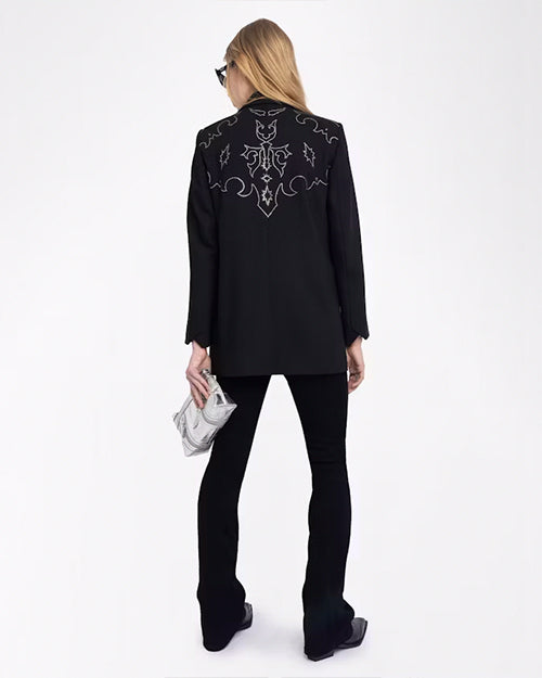 A full body backside view of a blonde model with her hair swept to the side wearing a black jacket with intricate Western style white embroidery at the top of the back and black pants to match. She is also wearing a silver clutch and black boots. The background is plain white.