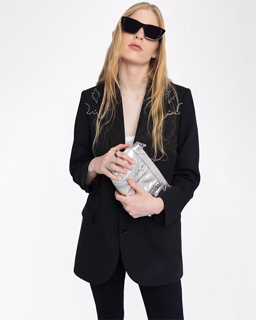 A knee-up view of a blonde model holding a silver clutch, wearing black pants with a black blazer featuring white embroidery at the shoulders, above a white shirt. She is also wearing black sunglasses and is standing in front of a white background.