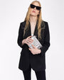 A knee-up view of a blonde model holding a silver clutch, wearing black pants with a black blazer featuring white embroidery at the shoulders, above a white shirt. She is also wearing black sunglasses and is standing in front of a white background.