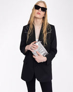 A knee-up view of a blonde model holding a silver clutch, wearing black pants with a black blazer featuring white embroidery at the shoulders, above a white shirt. She is also wearing black sunglasses and is standing in front of a white background.
