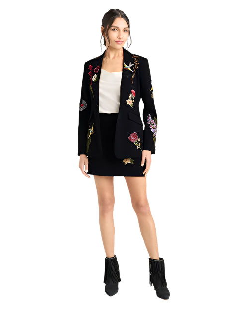 Woman wearing a black embroidered blazer and skirt with colorful designs on a white background