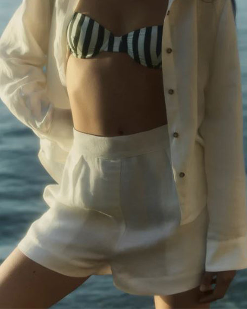 A person wearing white linen shorts with a matching top, shirt is open showing  a white and black striped swimsuit top.