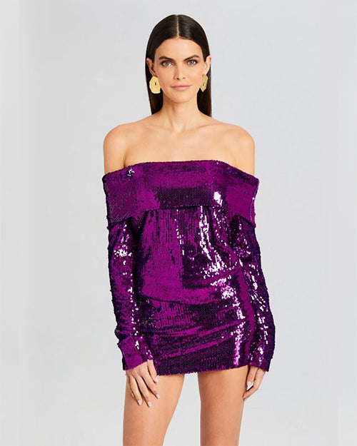 A close-up shot of a dark-haired model wearing a purple sparkly off-the-shoulder dress with long sleeves. The model is accessorizing with round gold earrings and standing in front of a white background.