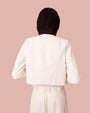 A back view of a model wearing a white jacket with a structured fit and purposeful topstitching. It has full-length sleeves and a standard collar. The model is styled with white pants.