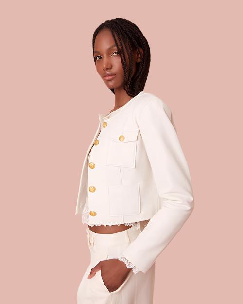 Side view of a model wearing a white jacket with gold buttons. The jacket features a structured design and includes four pockets on the front. The model wears matching high-waisted white pants.