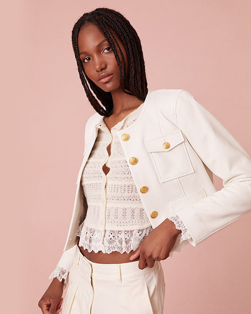 A knee-up frontal view of a brunette model wearing an open white jacket with gold buttons, a chest pocket and lace hems over a lacey white button-down shirt. The model is wearing matching white pants and is standing against a pink background.