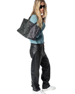A person with long hair wearing dark sunglasses, a blue shirt, black leather pants, and white sneakers, carrying a large black handbag with a patterned design on the shoulder.