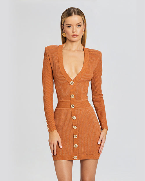A zoomed in image of a blonde model wearing an orange long sleeve dress with a ribbed material, a V-neck and gold buttons. The model is accessorizing with gold earrings and is standing in front of a white background.