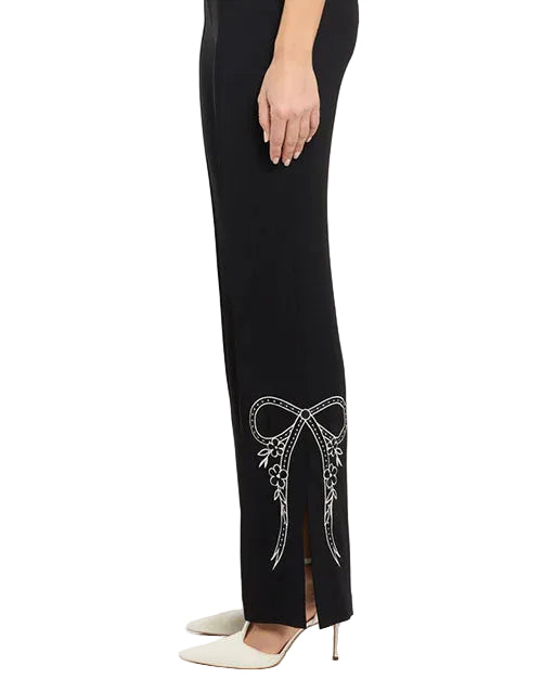 Side view of black tailored trousers with a slim fit and slight flare at the hem, highlighting white embroidered bow and floral detailing near the lower leg, worn with pointed-toe heels and photographed against a white background.