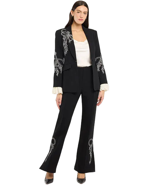Full-body studio view of a tailored black blazer and matching trousers set featuring white embroidered floral and bow detailing on the lapel, sleeves, and pant legs, layered over a white satin camisole and finished with flared trousers, photographed against a white background.