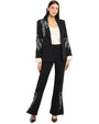 Full-body studio view of a tailored black blazer and matching trousers set featuring white embroidered floral and bow detailing on the lapel, sleeves, and pant legs, layered over a white satin camisole and finished with flared trousers, photographed against a white background.