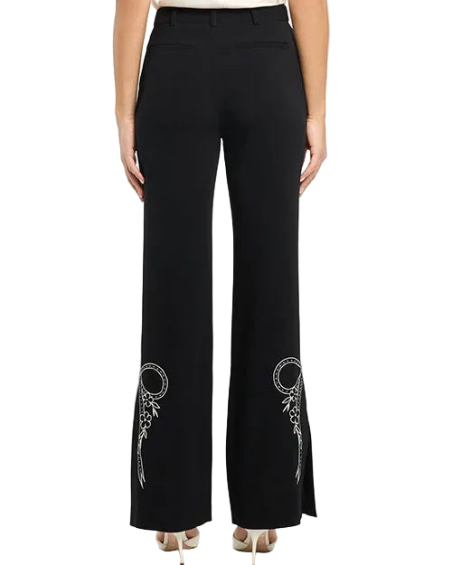 Back view of black tailored trousers with a straight-to-slightly flared leg, featuring white embroidered bow and floral detailing near the hems on both legs, paired with heels and photographed against a white background.