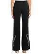 Back view of black tailored trousers with a straight-to-slightly flared leg, featuring white embroidered bow and floral detailing near the hems on both legs, paired with heels and photographed against a white background.