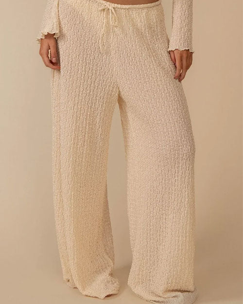 A person wearing loose, wide‑leg pants made from a cream-colored textured fabric with a nubby, boucle‑like surface. The pants have a relaxed fit and feature a drawstring waistband with ties at the front.