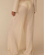 Back view of a person wearing loose, wide‑leg pants made from a cream-colored textured fabric with a nubby, boucle‑like surface. The pants have a relaxed fit and feature a drawstring waistband with ties at the front. 
