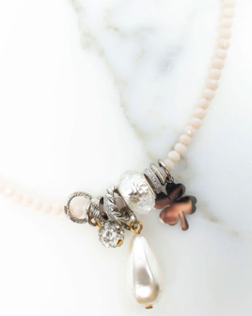 Close-up of a necklace with pale pink beads and a cluster of silver and metallic charms at the center, including a textured bead, a clear crystal, a teardrop-shaped pearl pendant, and a bronze four-leaf clover charm, displayed on a white marble surface.