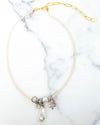 A necklace with small pale pink beads and a gold chain extender, featuring a cluster of metallic charms at the center, including a textured silver bead, a clear crystal, a bronze four-leaf clover, and a teardrop-shaped pearl pendant, displayed on a white marble surface.