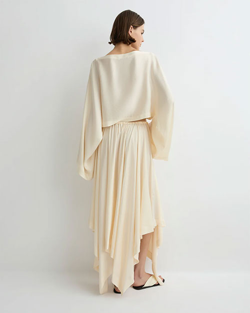 Back view of a person wearing a soft cream long-sleeve cropped top with wide draped sleeves and a matching flowing midi skirt featuring an asymmetrical hem, paired with light-colored flat sandals against a plain light background.
