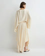 Back view of a person wearing a soft cream long-sleeve cropped top with wide draped sleeves and a matching flowing midi skirt featuring an asymmetrical hem, paired with light-colored flat sandals against a plain light background.