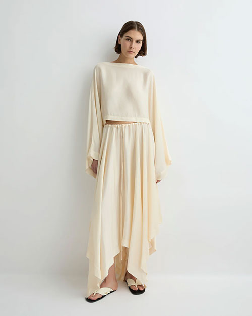 Person modeling a soft cream long-sleeve cropped top with a round neckline and matching flowing midi skirt, paired with light-colored flat sandals against a plain light background.