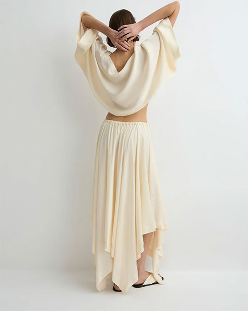 Rear view of a person modeling a soft cream long-sleeve cropped top with a round neckline and matching flowing midi skirt, paired with light-colored flat sandals against a plain light background.