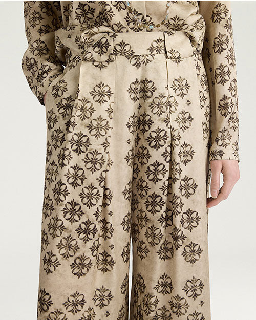 Close-up view of cream-colored wide-leg trousers featuring an allover black floral-inspired geometric print. The high-waisted trousers have front pleats, a matching belt, and a relaxed, flowing silhouette. Shown styled with a coordinating printed top against a light gray background.