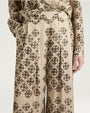 Close-up view of cream-colored wide-leg trousers featuring an allover black floral-inspired geometric print. The high-waisted trousers have front pleats, a matching belt, and a relaxed, flowing silhouette. Shown styled with a coordinating printed top against a light gray background.